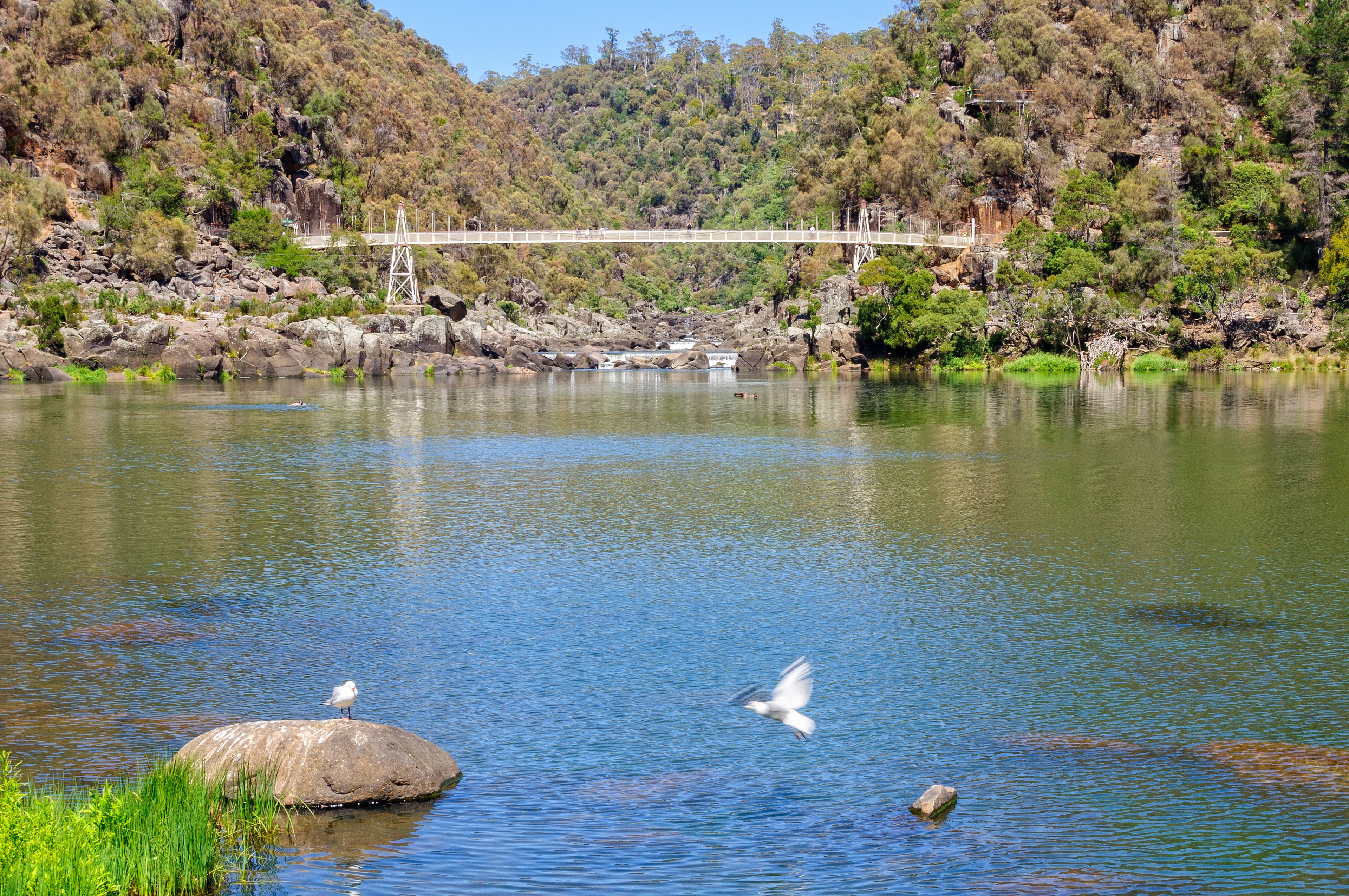 A calm river with a bridge and rocky hills in the background.