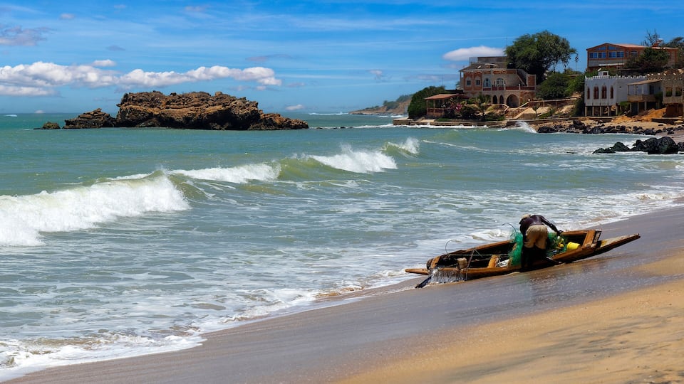 A beach with waves, a rocky outcrop, and houses in the background.