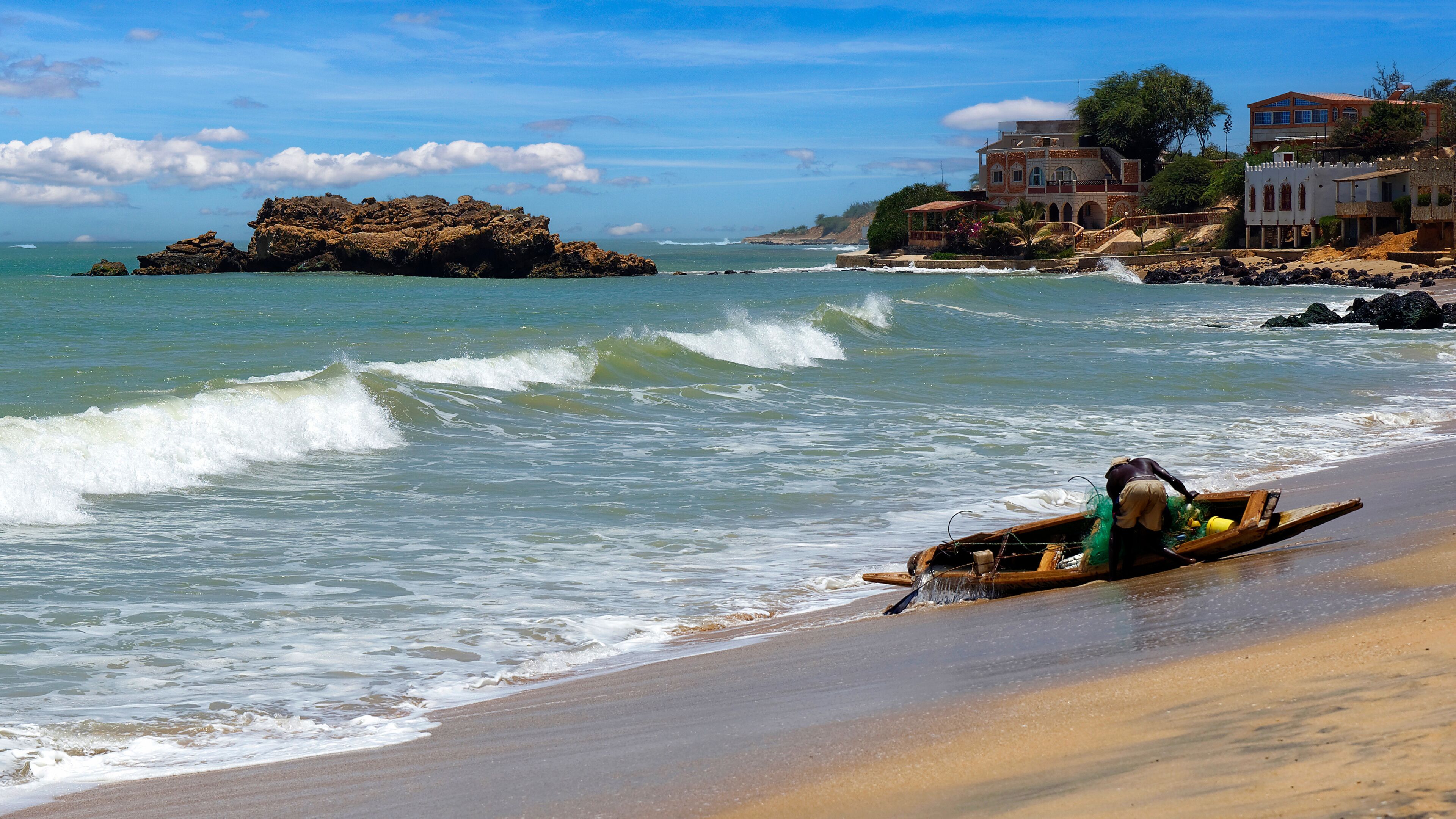 A beach with waves, a rocky outcrop, and houses in the background.