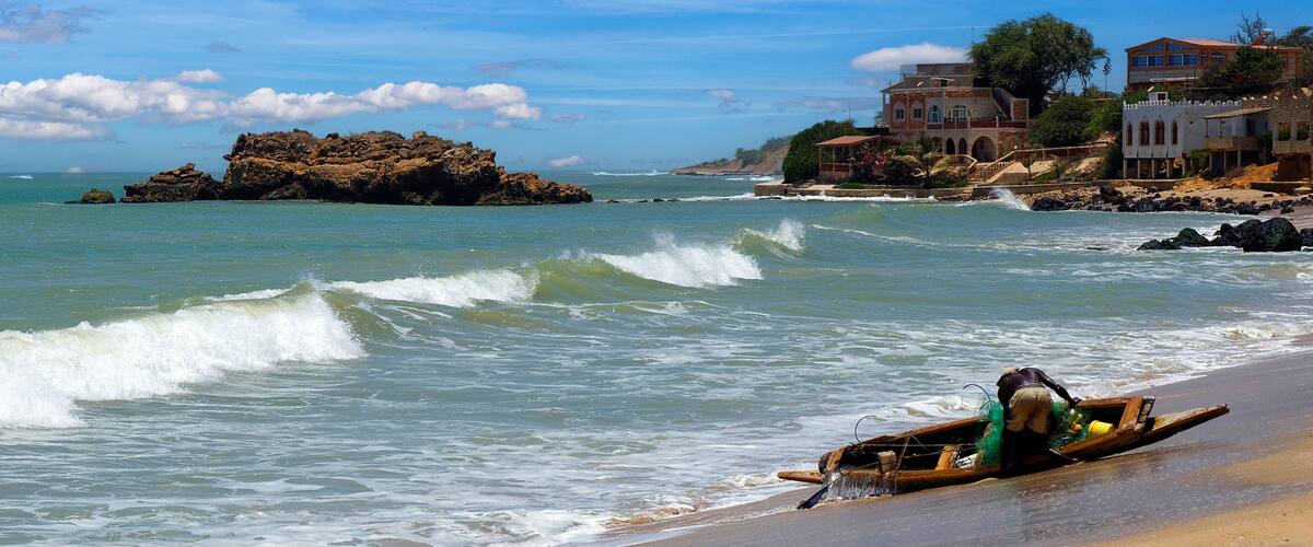 A beach with waves, a rocky outcrop, and houses in the background.