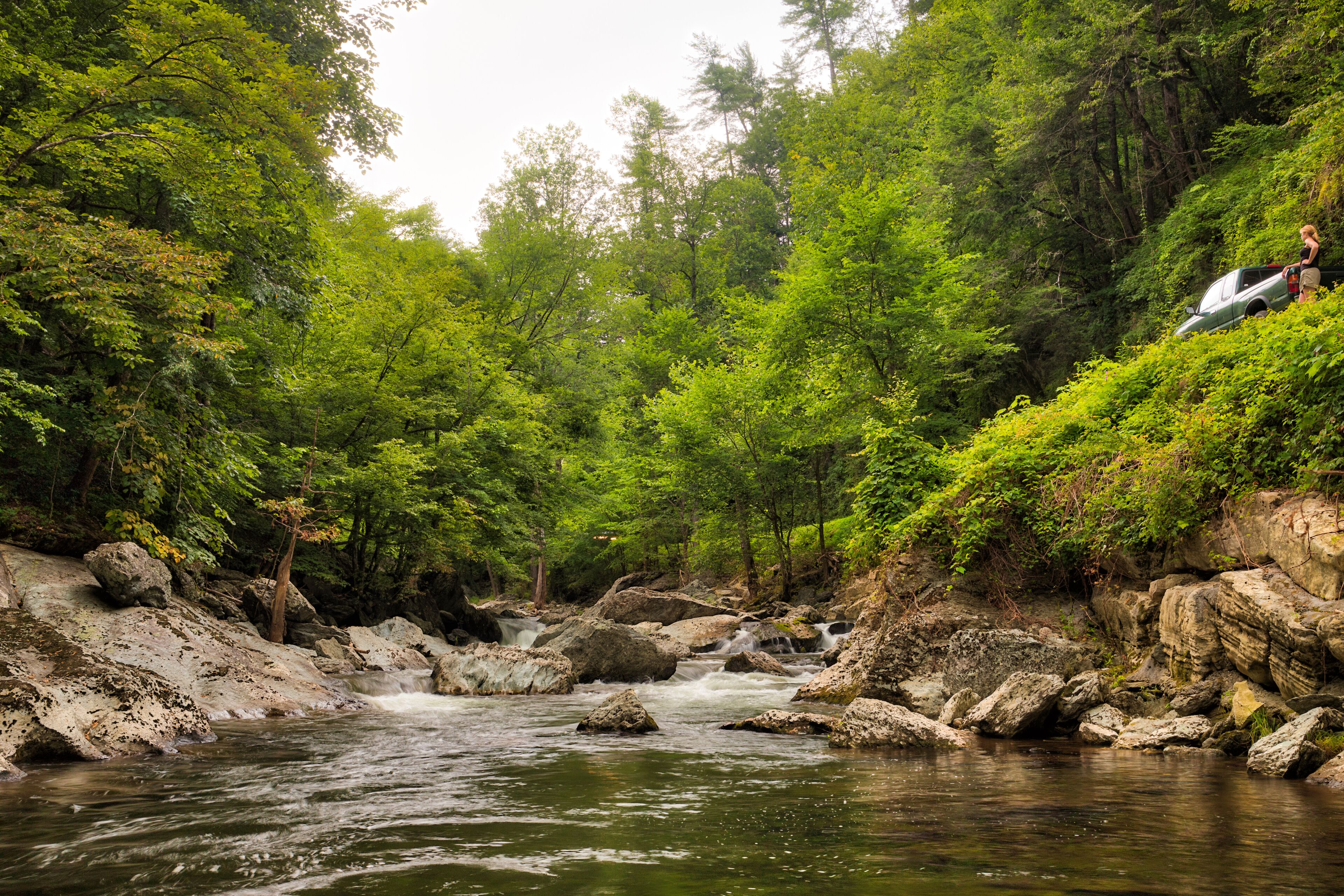 River and green trees in the Smoky Mountains with a caucasian woman parked beside it overlooking it next to her pickup truck