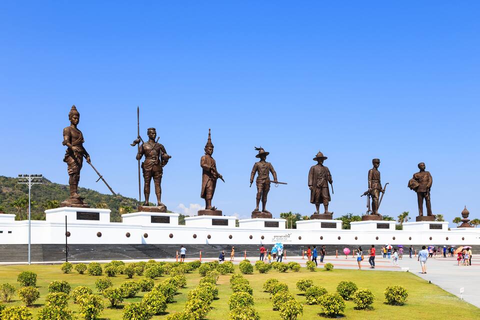 A row of bronze statues on pedestals, a white building with a sign, and a well-maintained grassy area with bushes.