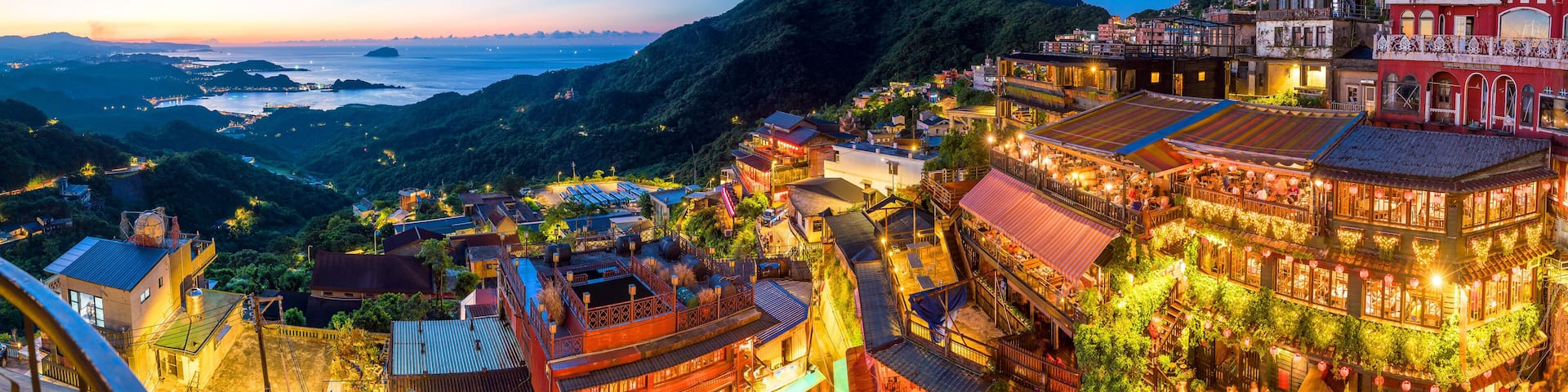 A coastal town at dusk with illuminated buildings, a prominent mountain, and a view of the sea.