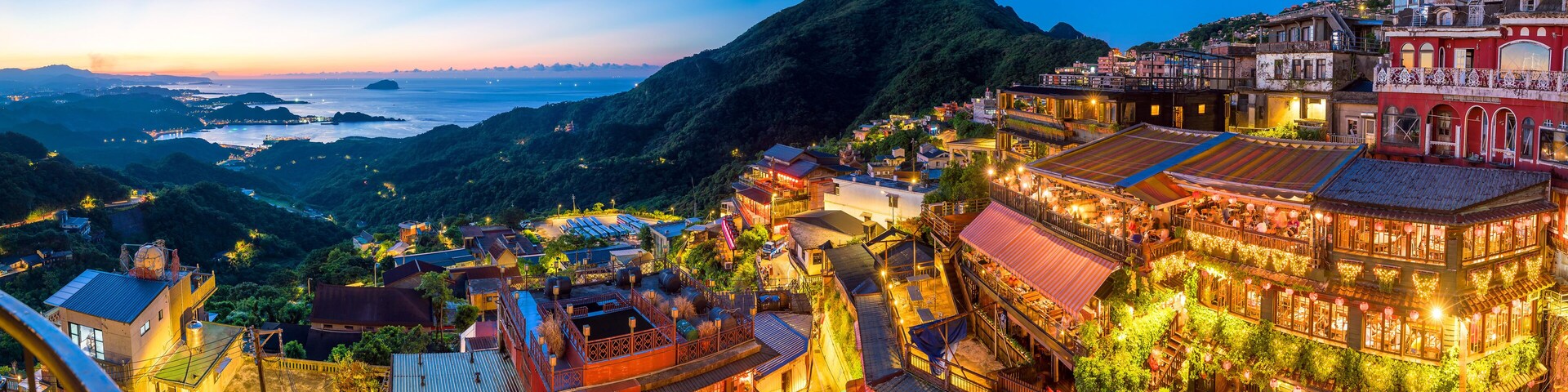 Uma vila costeira ao entardecer, com edifícios iluminados, uma montanha imponente e vista para o mar.