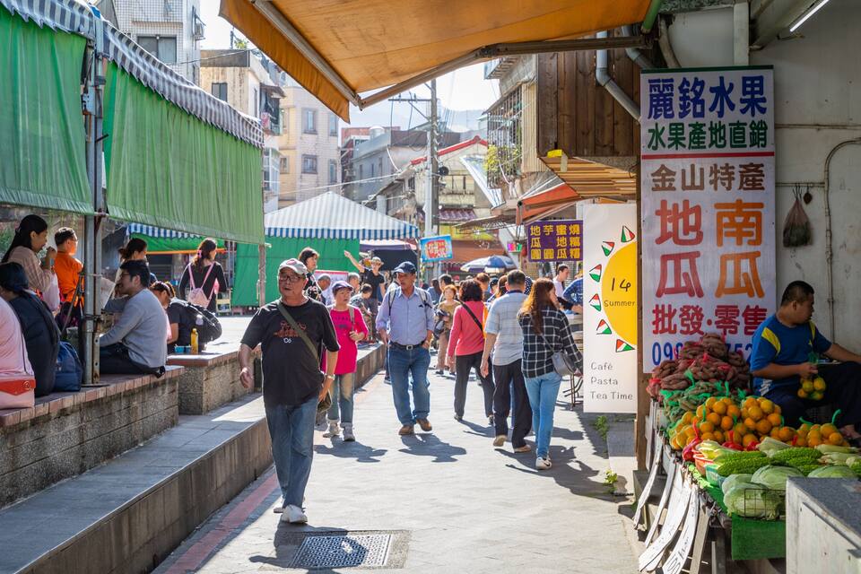 Um mercado de rua movimentado, com pessoas caminhando e vendedores oferecendo frutas e legumes.