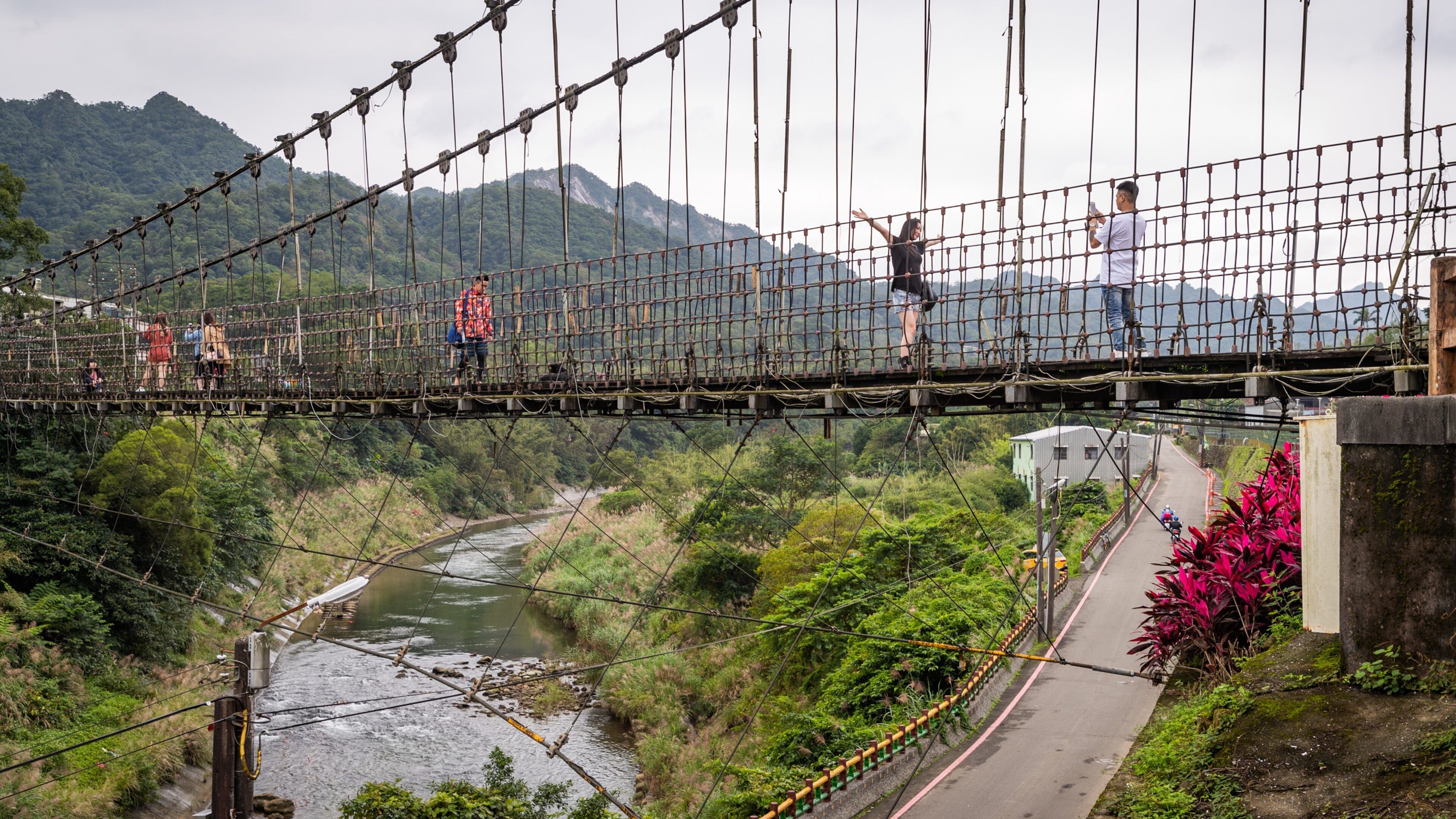 A suspension bridge over a river with lush greenery and mountains in the background.