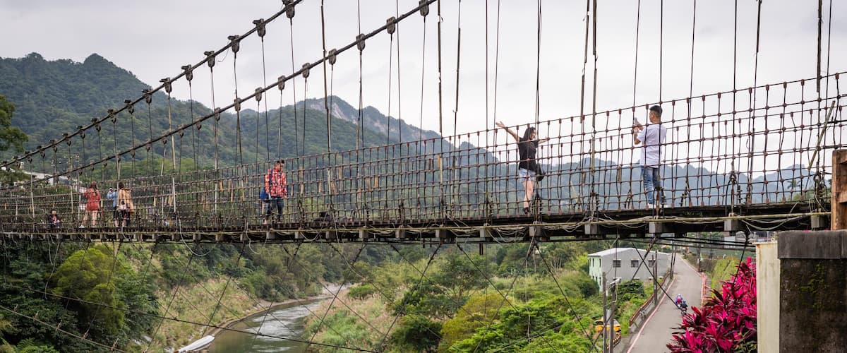 A suspension bridge over a river with lush greenery and mountains in the background.