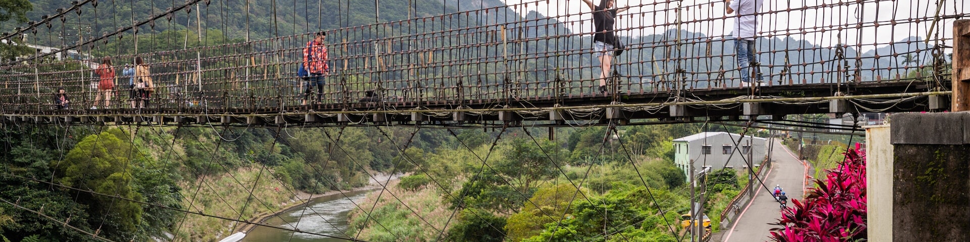 A suspension bridge over a river with lush greenery and mountains in the background.