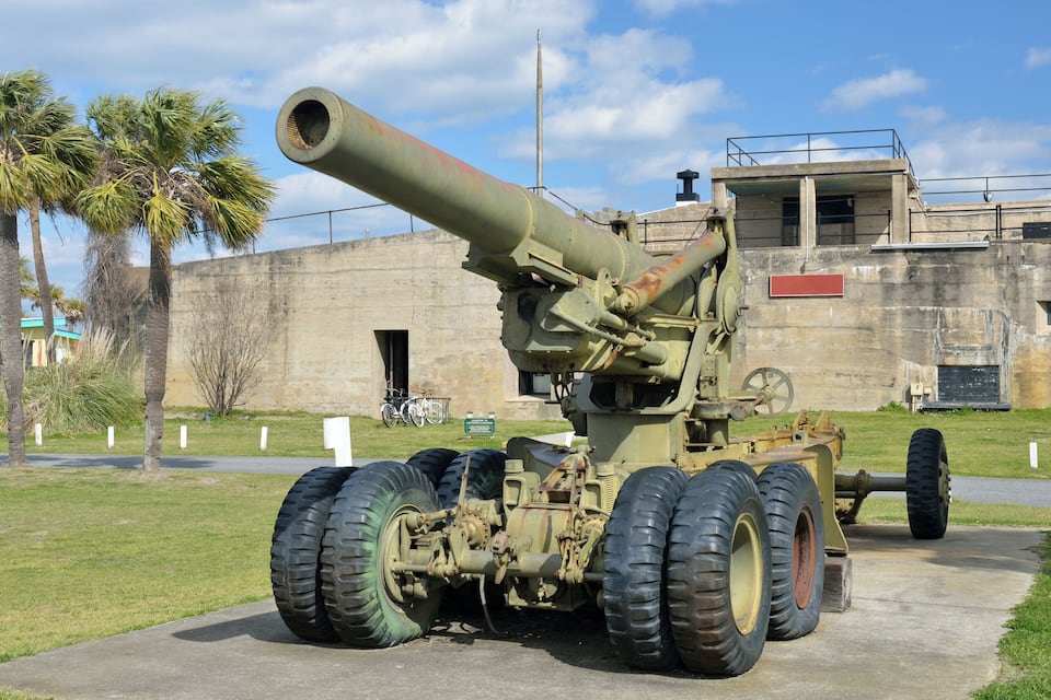 A large artillery piece with large wheels, parked in front of a building with palm trees and a clear sky.