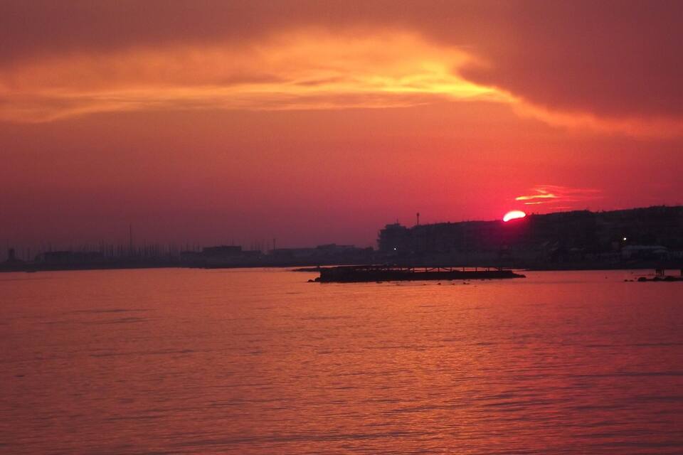 Um pôr do sol sobre a água, com o horizonte de uma cidade silhuetado ao fundo.