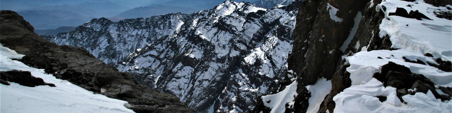 A snow-covered mountain range with rocky peaks and a cloudy sky.
