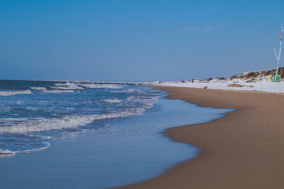 Uma praia com ondas, céu claro e horizonte urbano ao longe.