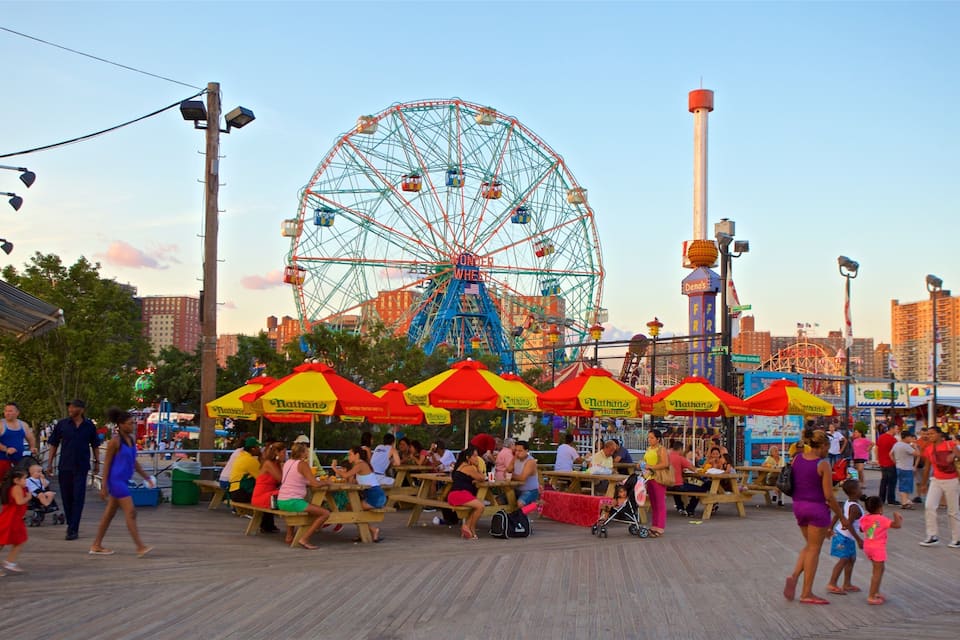A boardwalk with picnic tables and umbrellas, a Ferris wheel, and a food vendor named Nathan's in the background.
