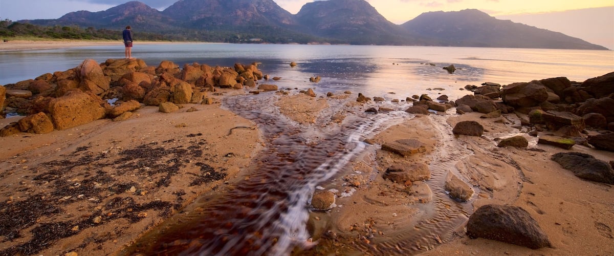 A rocky beach with a person standing on the rocks, a flowing stream, and mountains in the background.