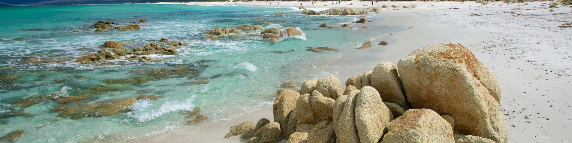 A beach with clear turquoise waters, large rocks in the foreground, and a hilly landscape in the background.