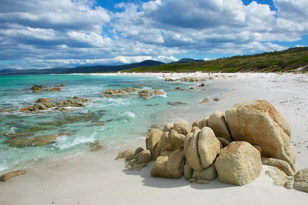 A beach with clear turquoise waters, large rocks in the foreground, and a hilly landscape in the background.