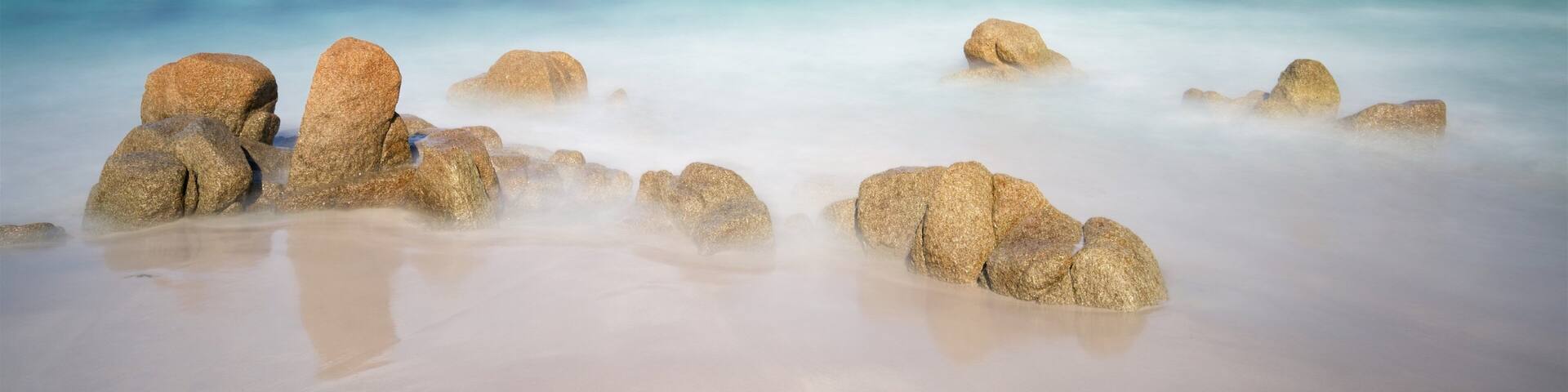 Una playa con rocas grandes al frente y un mar tranquilo bajo un cielo nublado.
