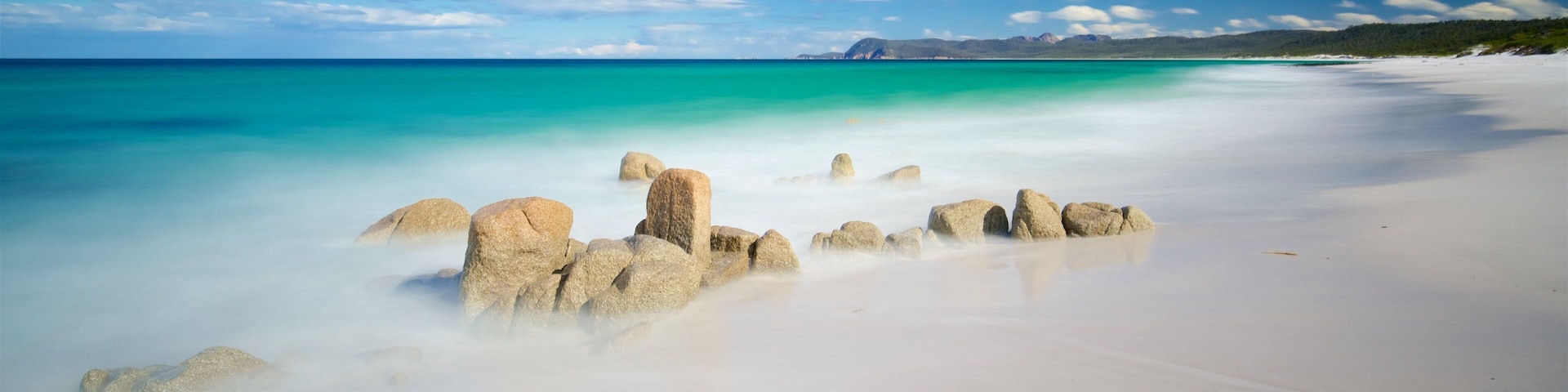 Una playa con arena blanca y agua turquesa, con rocas grandes al frente y un cielo despejado con nubes.