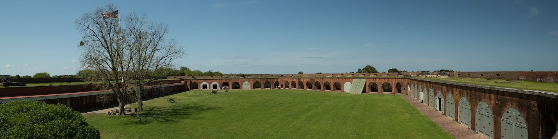 A large, open courtyard with a grassy lawn, brick buildings with arched doorways, and a flagpole with an American flag.