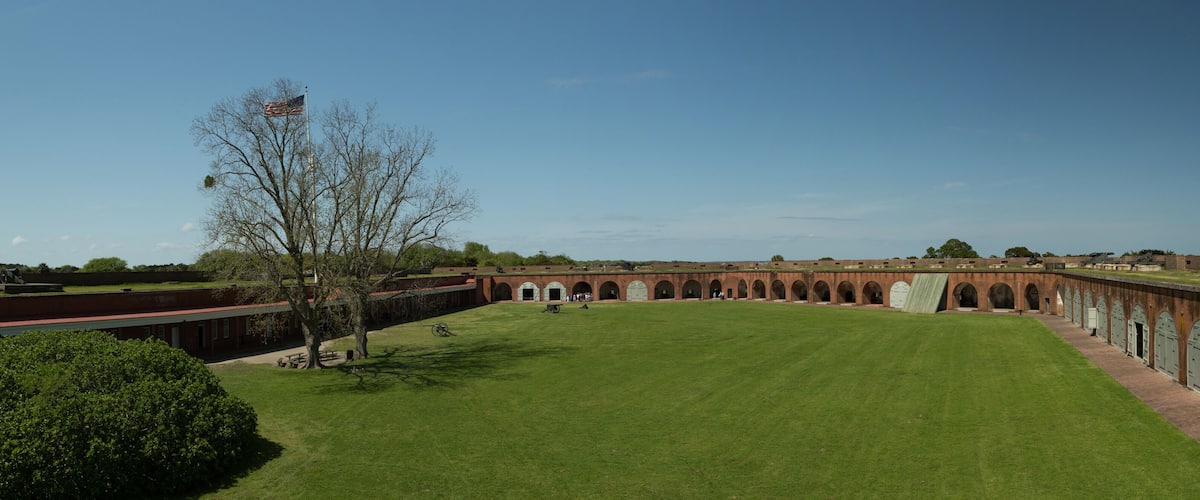 A large, open courtyard with a grassy lawn, brick buildings with arched doorways, and a flagpole with an American flag.