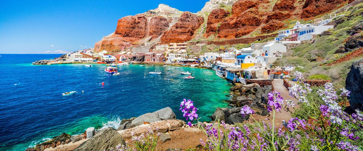 A coastal village with white buildings, clear blue sea, rocky cliffs, and purple flowers.