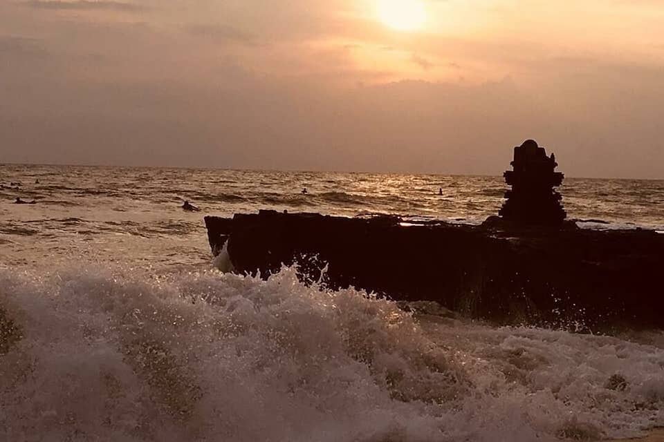 A sunset over the ocean with waves crashing against a rocky outcrop.