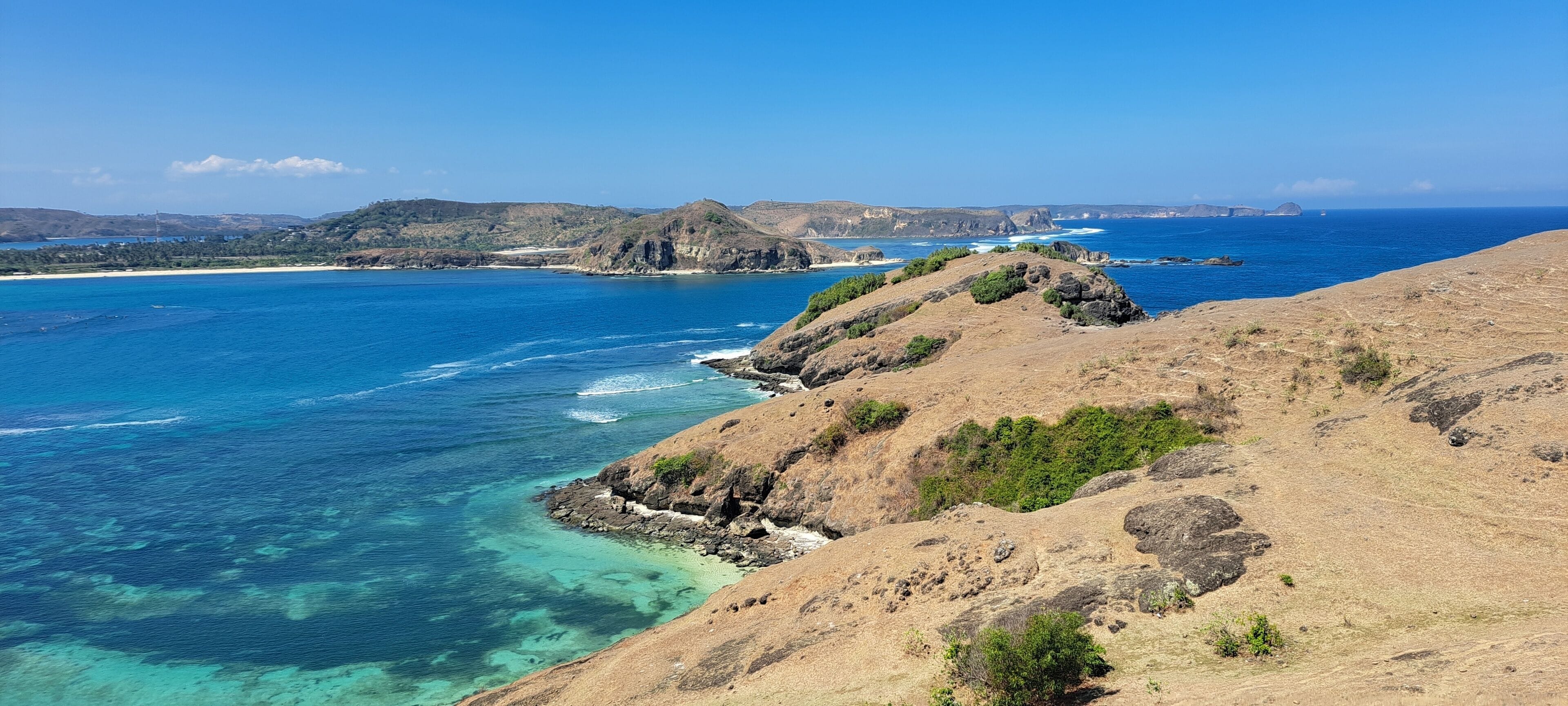 Paisagem costeira com águas azul-claras, falésias rochosas e colinas ao longe.