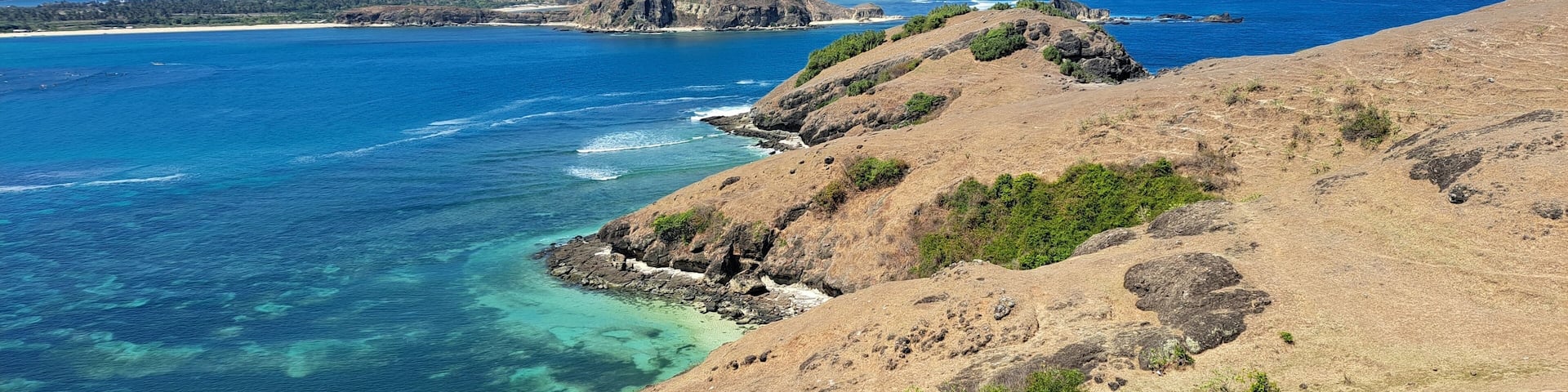 A coastal landscape with clear blue waters, rocky cliffs, and distant hills.