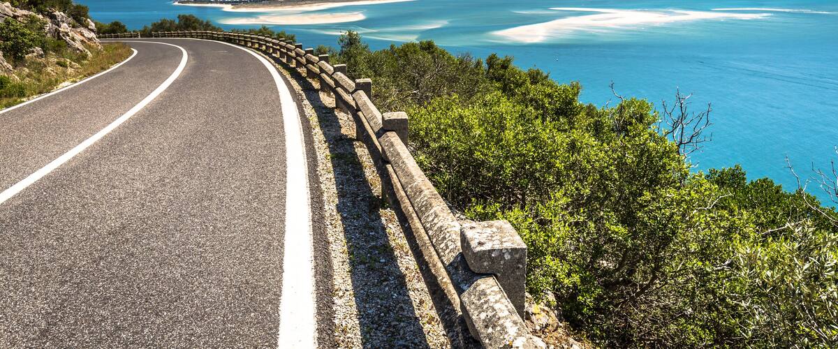 A curving road with a guardrail, overlooking a coastal area with clear blue waters and distant buildings.