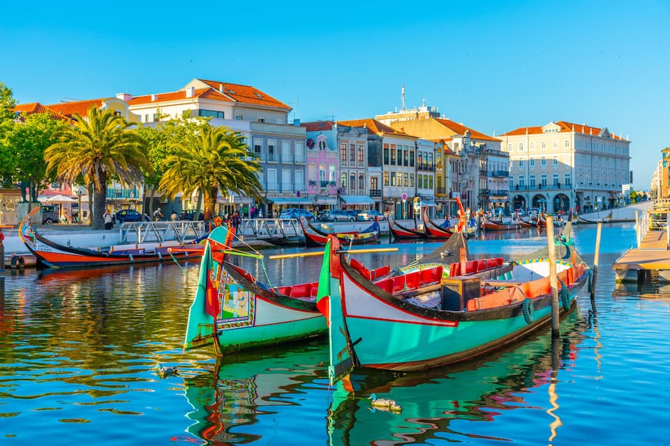 Um porto com barcos coloridos atracados, palmeiras e edifícios históricos com telhados vermelhos.