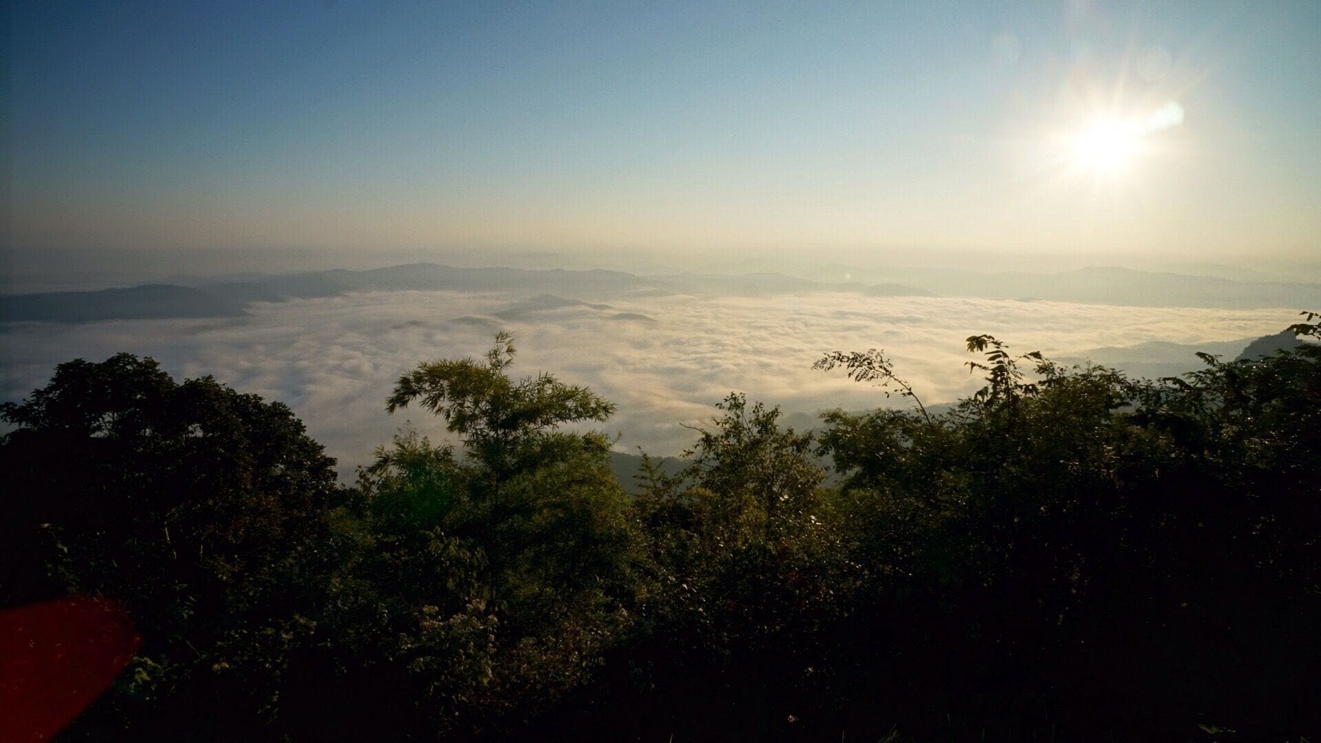 A mountain landscape with a view of the clouds below and the sun shining in the sky.