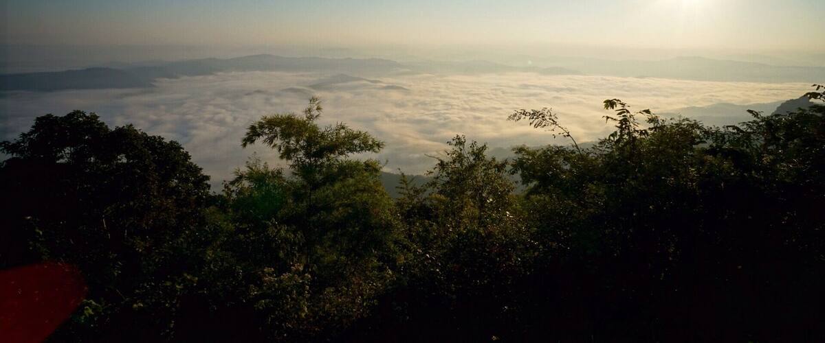 A mountain landscape with a view of the clouds below and the sun shining in the sky.