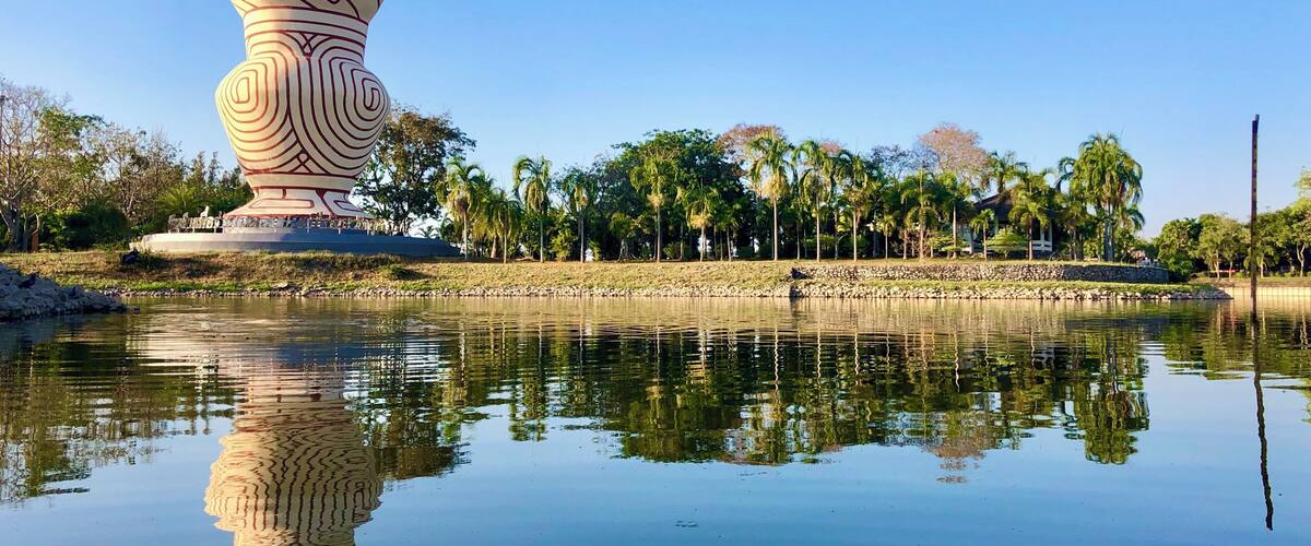 A large, uniquely shaped sculpture by a calm body of water with palm trees and a clear blue sky in the background.