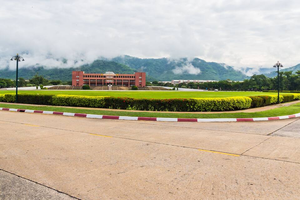 A large building with a red facade, surrounded by well-maintained green lawns and hedges, with mountains in the background.