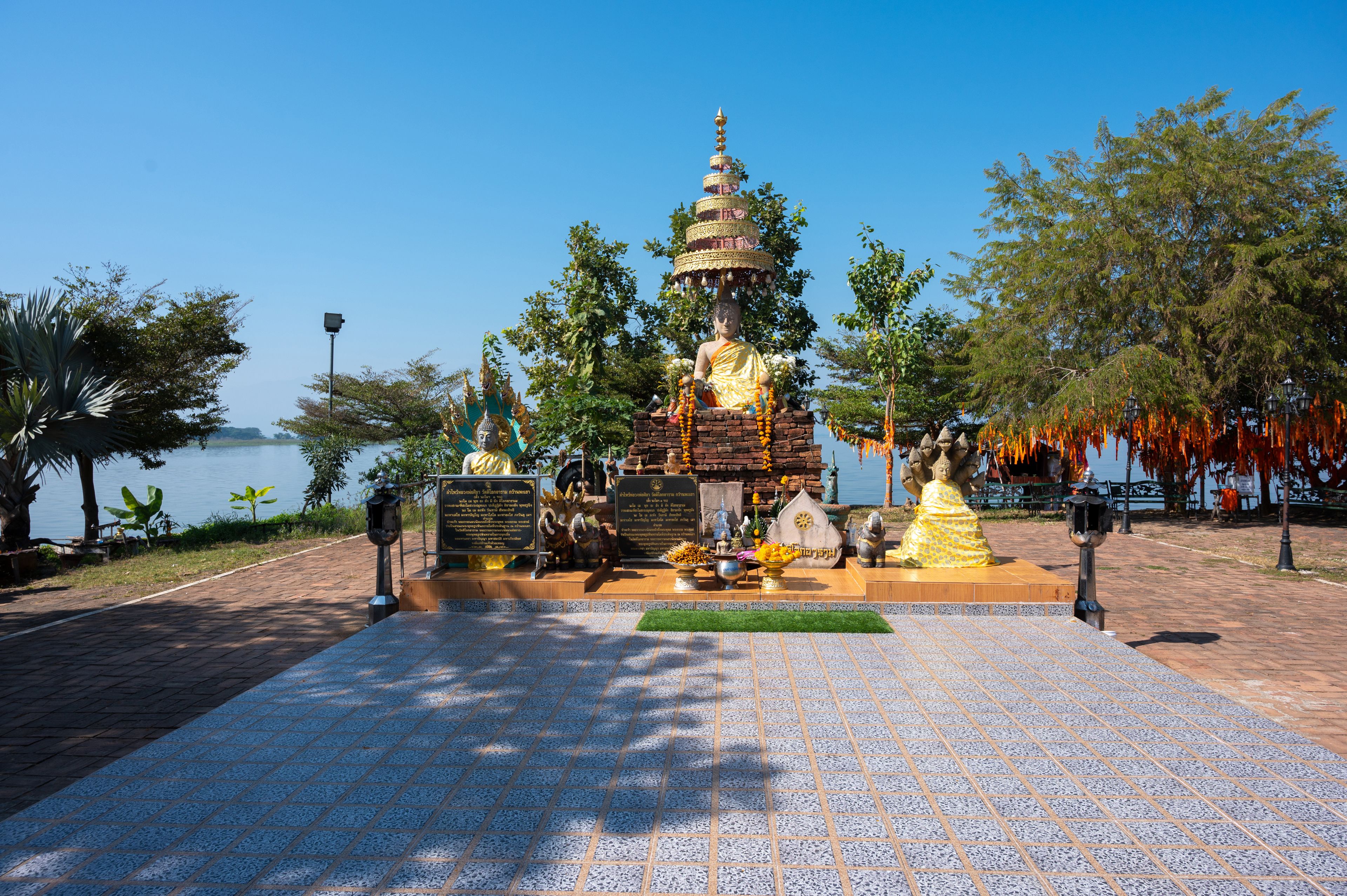 A temple with a large Buddha statue, surrounded by smaller statues and offerings, set against a backdrop of trees and a body of water.