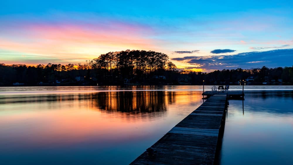 Un lac paisible au crépuscule, avec une jetée en bois qui s’avance sur l’eau, entouré d’arbres et sous un ciel dégagé.