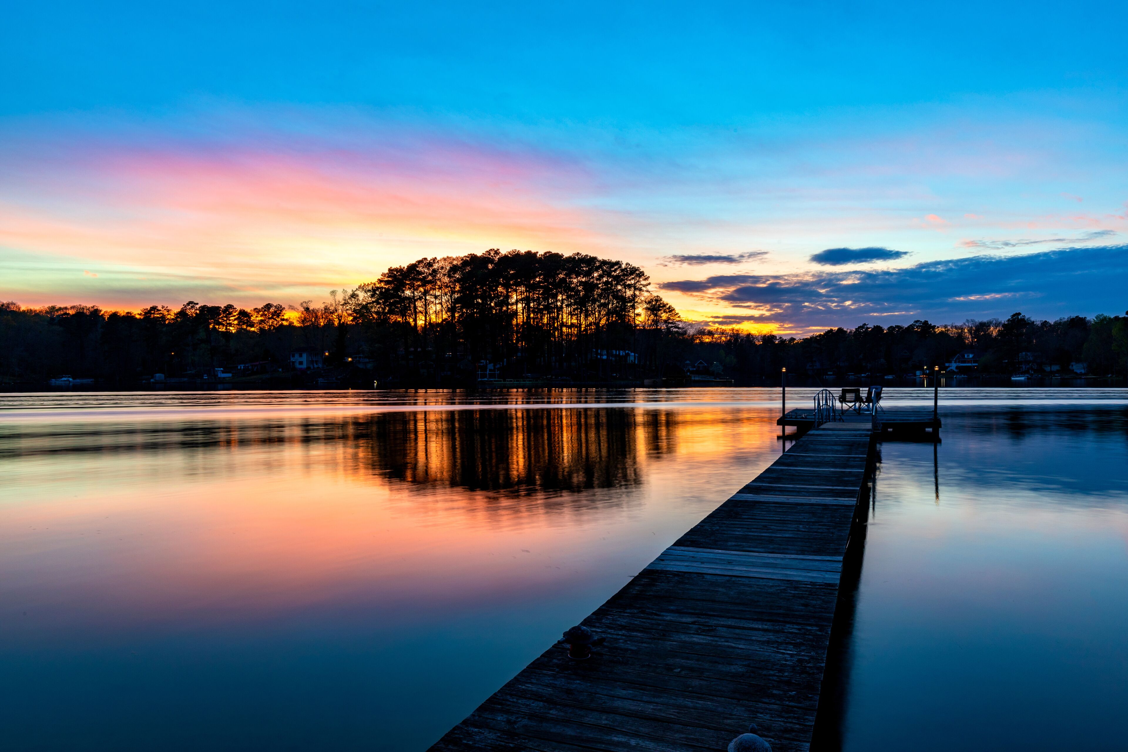 A calm lake at dusk with a wooden dock extending into the water, surrounded by trees and a clear sky.