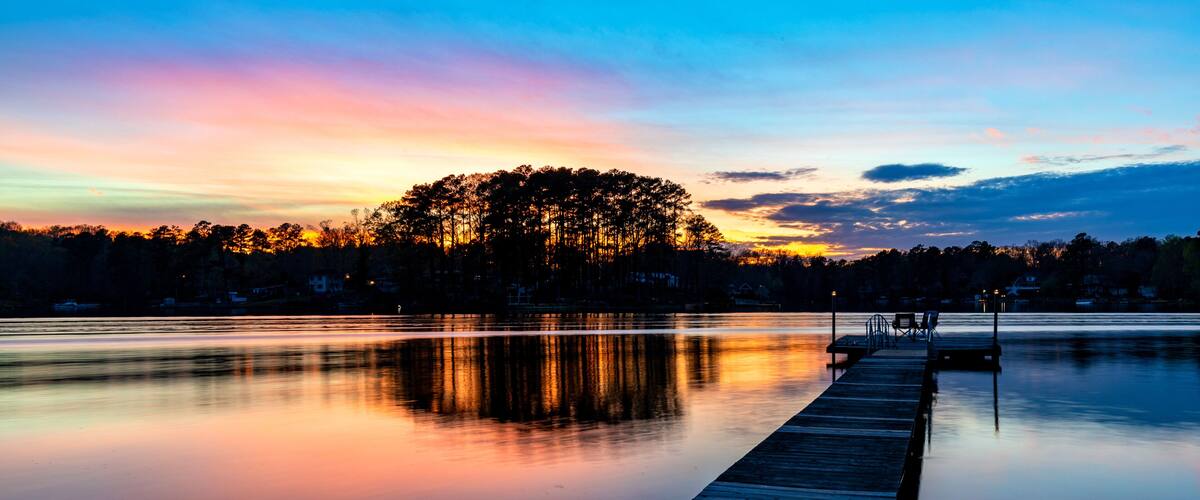 A calm lake at dusk with a wooden dock extending into the water, surrounded by trees and a clear sky.