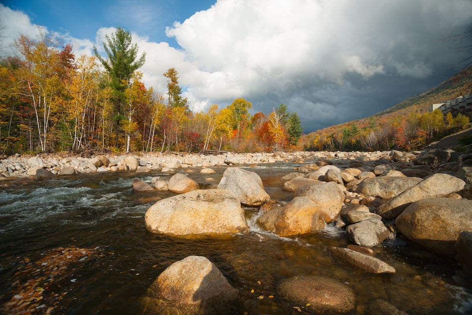 A river with large rocks, autumn-colored trees, and a cloudy sky.