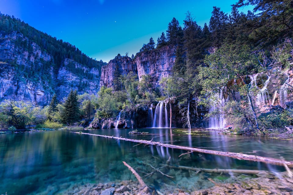 Um lago tranquilo com uma cascata, cercado por uma floresta densa e penhascos rochosos.