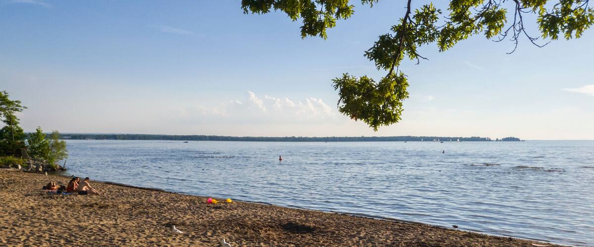 A sandy beach with a wooden fence, a calm sea, and a distant shoreline with sailboats.