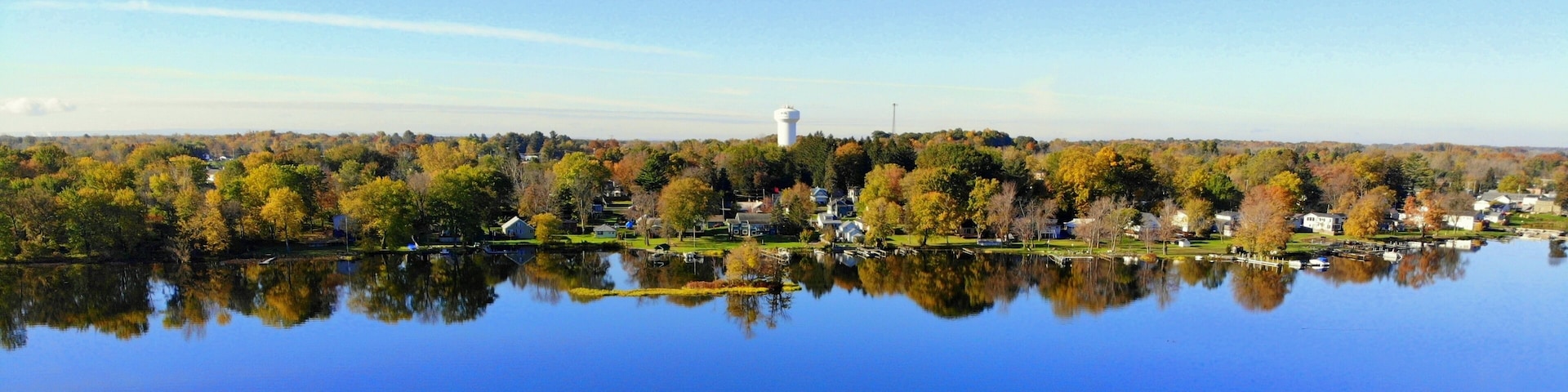 Ein ruhiger See mit Spiegelbild von Bäumen und Häusern, ein klarer Himmel und ein ferner Wasserturm.