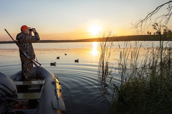 hunter looking through binoculars at sunset. waterfowl hunter on boat at lake. banner with place text.