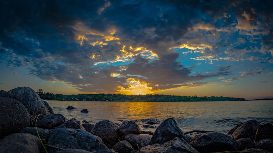 A sunset over a lake with rocks in the foreground and a forested shoreline.
