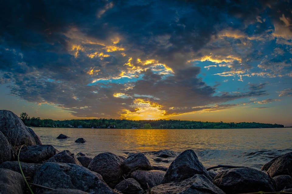 A sunset over a lake with rocks in the foreground and a forested shoreline.