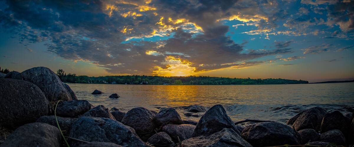 A sunset over a lake with rocks in the foreground and a forested shoreline.