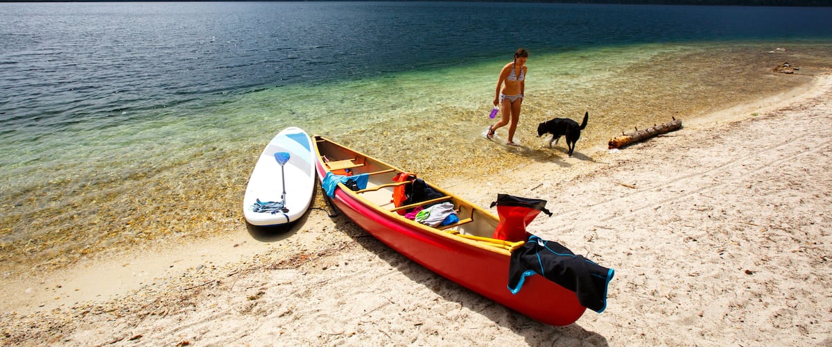 A lake with a red canoe and a surfboard on the shore, a dog, and a hiker in the background.