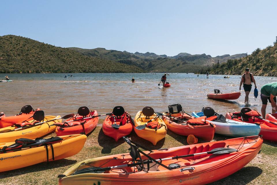 A group of kayaks lined up on the shore of a lake with people paddling in the water.