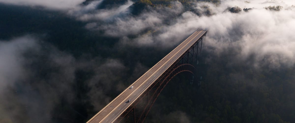 A bridge with a steel structure and a road, surrounded by clouds and a forested landscape.