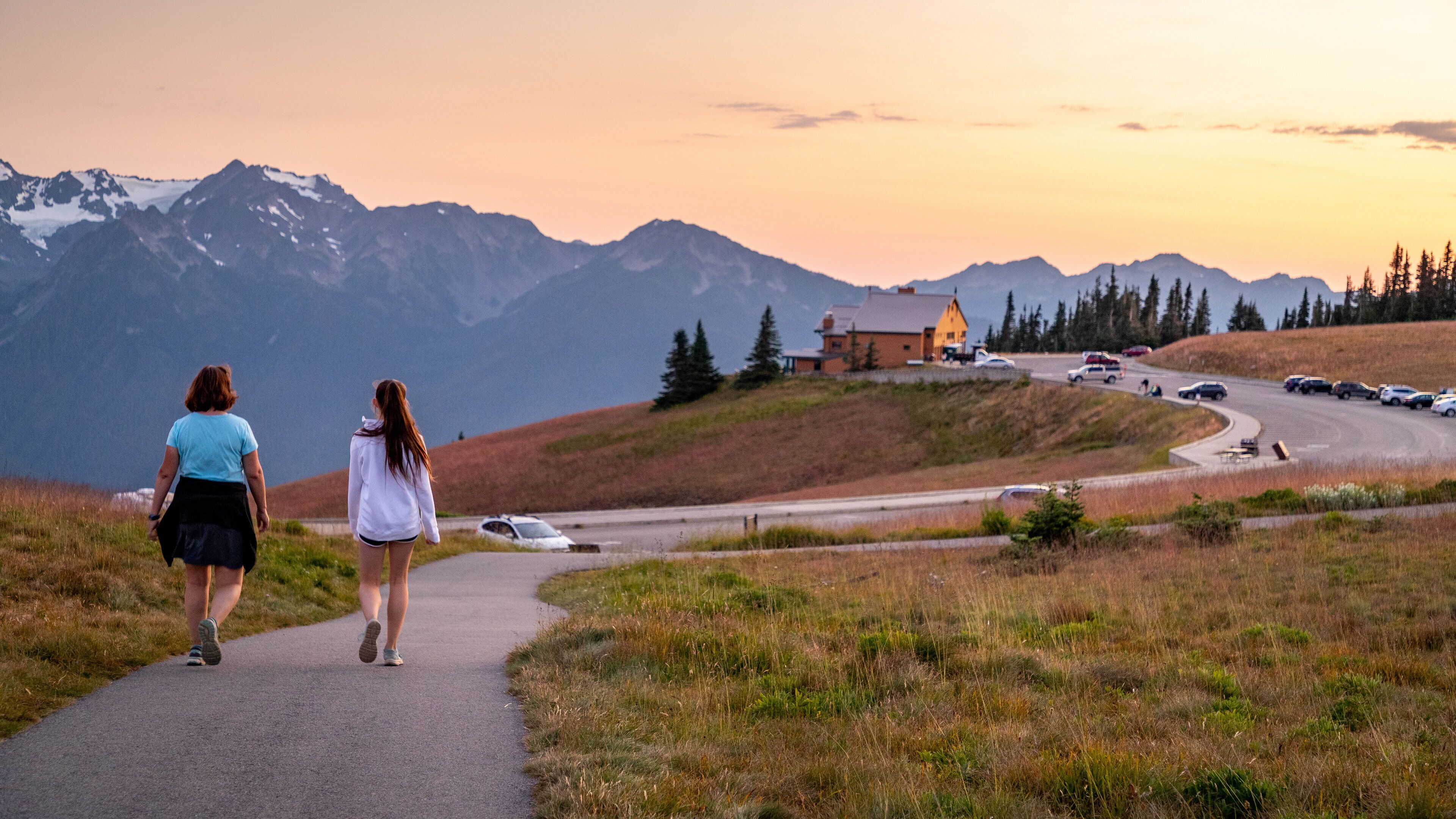 A paved path with two people walking, surrounded by mountains and a building with a yellow roof.